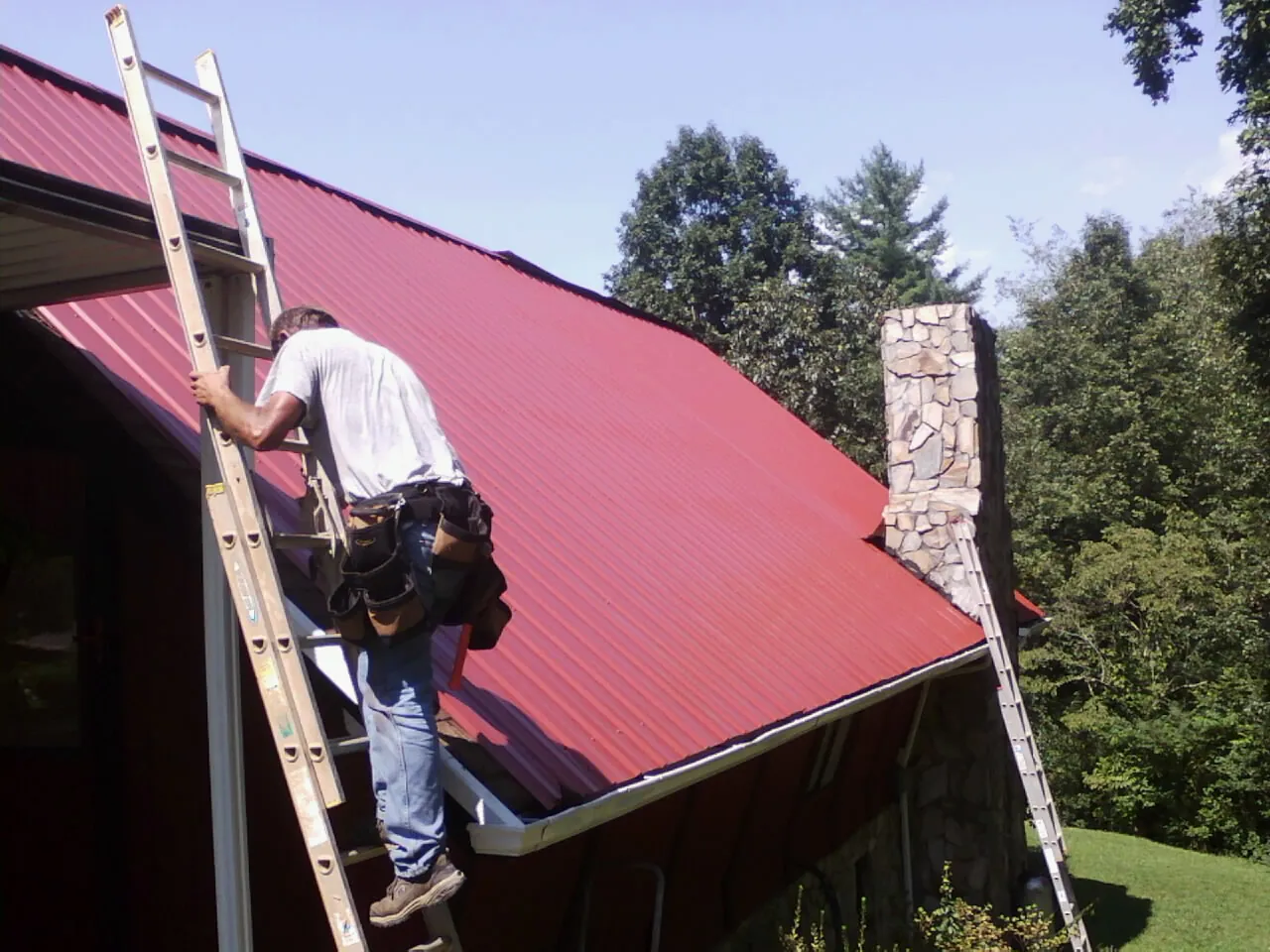 Roofer on a ladder beside a red metal roof and stone chimney — Soffit & Fascia Repair by Roofing Brothers in Pearl River
